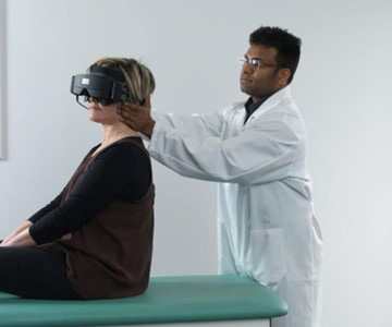 Clinician stood behind patient sat on a green examination bed. The clinician is turning the patient's head toward the left at a 45-degree angle.