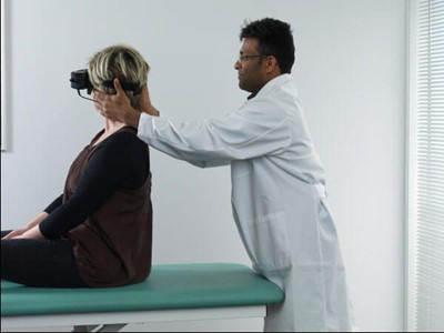 Clinician stood behind patient sat on a green examination bed. The clinician is turning the patient's head toward the right at a 45-degree angle.