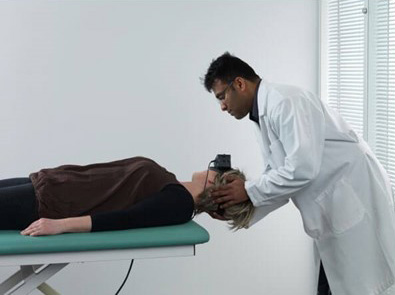 Clinician stood behind patient lying on their back on a green examination bed. The clinician is turning the patient's head toward the right at a 45-degree angle.