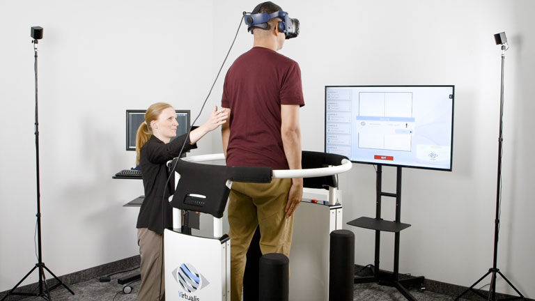 The patient is facing a monitor showing the SOT software. A clinician is stood next to the patient with their hand at a small distance behind the patient&rsquo;s back to offer support if needed.