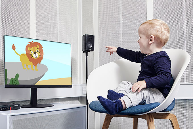 Young boy sat in a white chair on a blue cushion inside a booth. He&acirc;&euro;&trade;s looking and pointing toward a computer screen to his right that is showing a smiling animated lion. The VRA Screen computer and a speaker are located on each side of the computer screen.