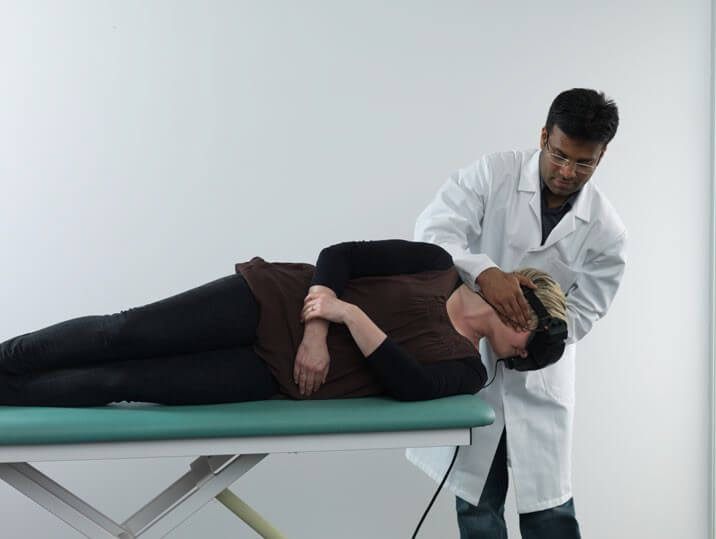 Female patient lying on her left side on examination table, with head hanging over the edge of the table. Male clinician is turning the patient's head toward the left, so it is pointing toward the floor.
