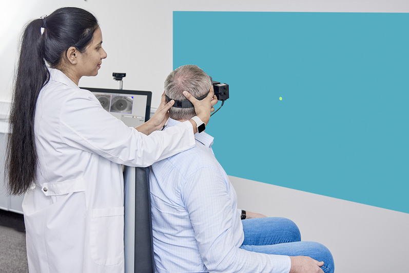 Clinician performing Gaze test in a patient. The patient is sat in the Orion Reclining rotary chair and fixated with VisualEyes VNG goggles, looking toward a dot projected onto a wall. Their body is facing 45 degrees to the right of the stimulus, and the clinician is turning the patients head 45 degrees to the left to look at the stimulus. The positioning is the same for the Smooth Pursuit Neck Torsion test.